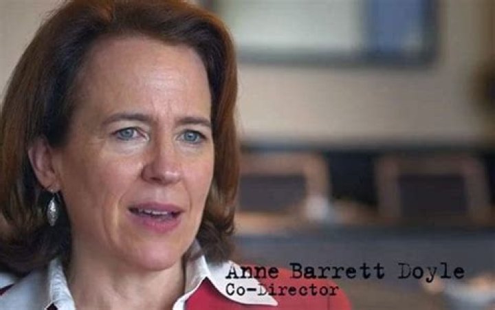 anne barrett doyle from the waist up in a black blouse standing in a stairwell next to a stained glass window leaving shadows on her face she has auburn hair, pink lipstick, and is 62 years old